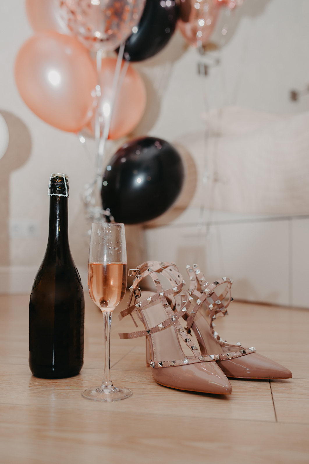invitation card for a bachelorette party, stylish beige sandals with spikes, a glass of pink champagne and a bottle on a background of pink and black balloons horizontally